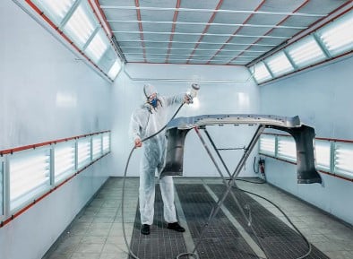 man in an enclosed room in full protective gear spray painting a piece of metal